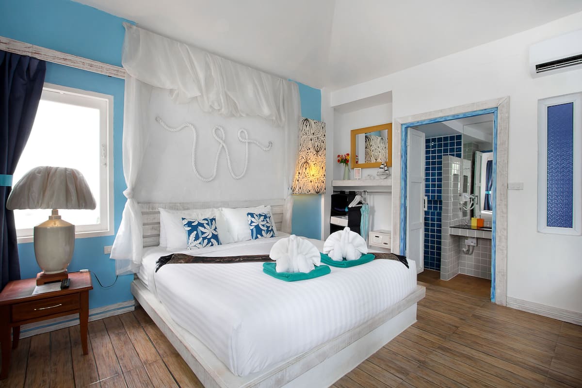 A bedroom with a bed, a nightstand, a lamp, a window with curtains, a bathroom visible through an open door, and a mirror on the wall.