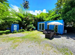 Un área de estar azul al aire libre con una mesa y sillas, rodeada de abundante follaje y una palmera.