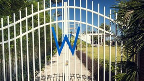 A gated entrance with a large blue 'W' sign, surrounded by greenery and a pathway leading to a building.