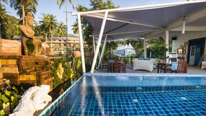 A swimming pool with a tiled edge, a statue, and a covered seating area with tables and chairs.