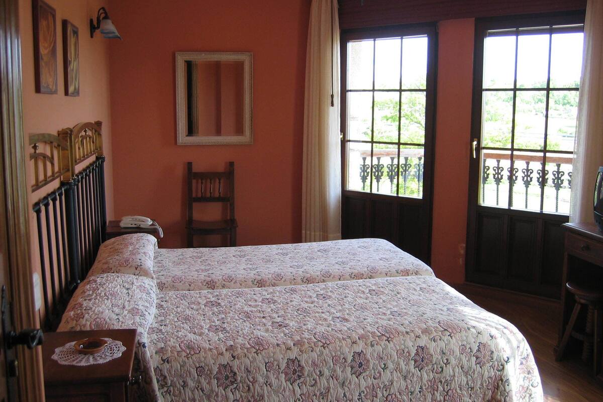 A bedroom with a double bed, a chair, a desk, a mirror, and a window with a view of greenery.