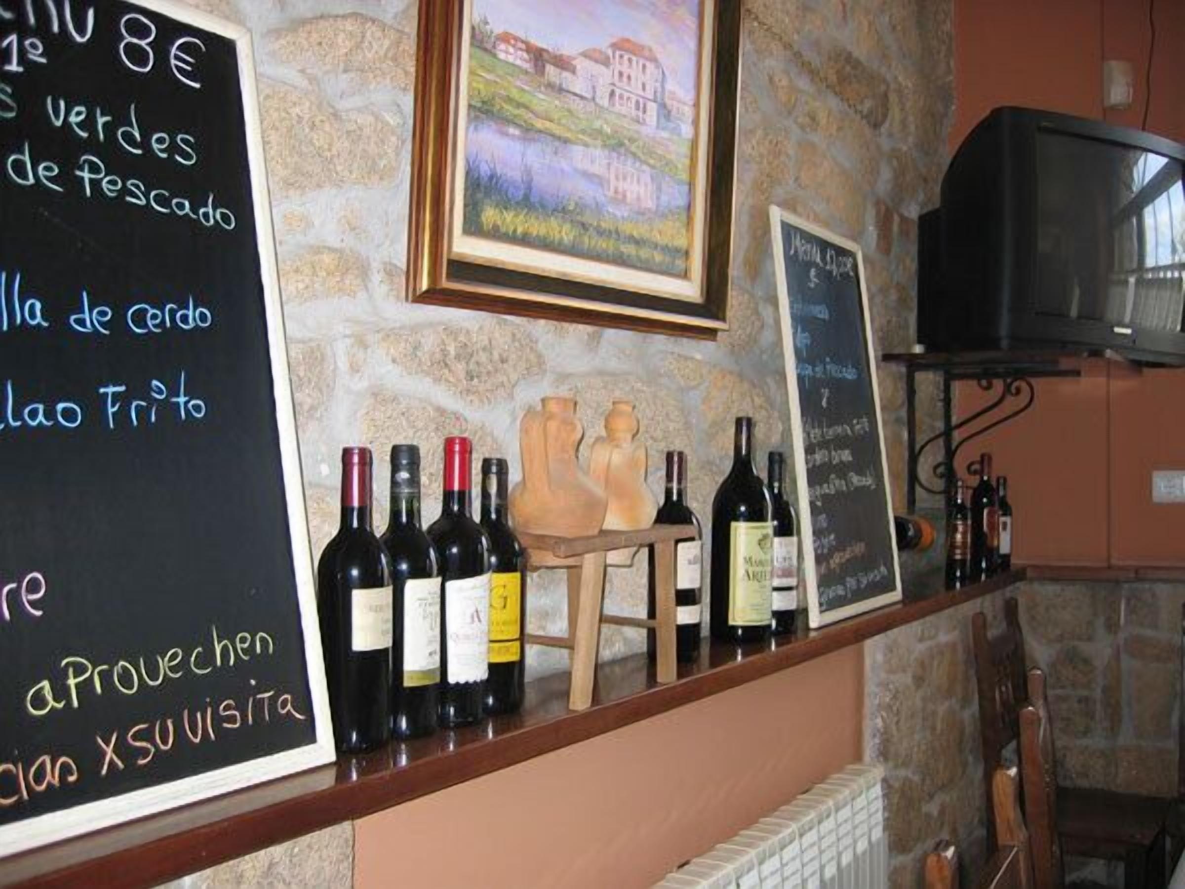 Zona de bar con un menú en pizarra que incluye opciones como 'Verde de Pescado' y 'a Provençale XSuvista'. Hay botellas de vino en la barra, una escultura de madera y un cuadro en la pared.