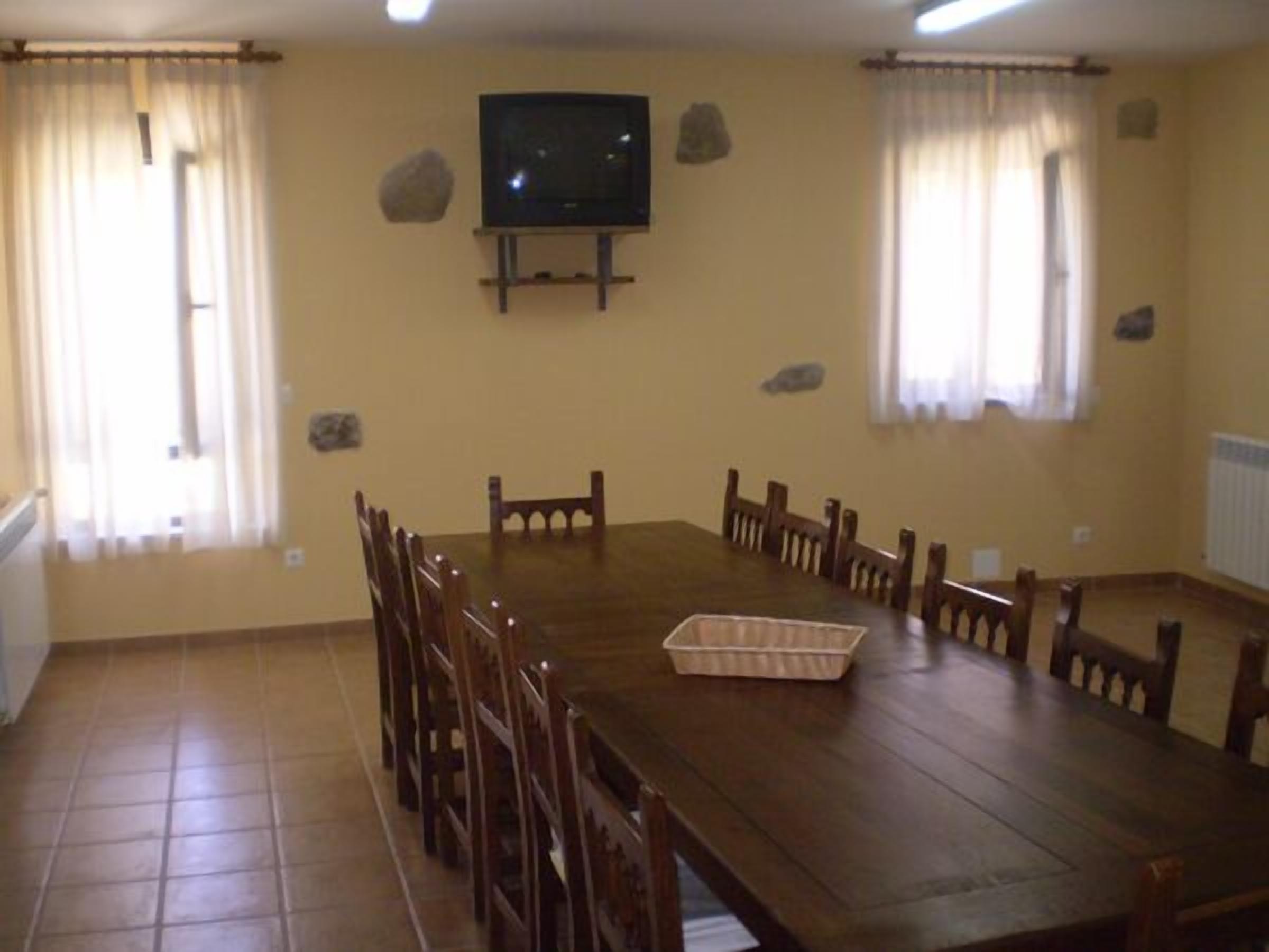 Un comedor con una gran mesa de madera, varias sillas, un televisor montado en la pared y dos ventanas con cortinas.