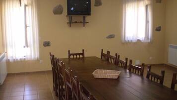 Un comedor con una gran mesa de madera, varias sillas, un televisor montado en la pared y dos ventanas con cortinas.