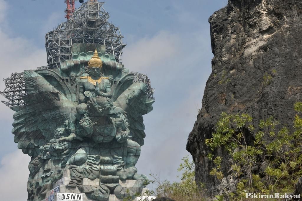 A large, ornate statue with intricate carvings and a golden top, set against a rocky cliff.