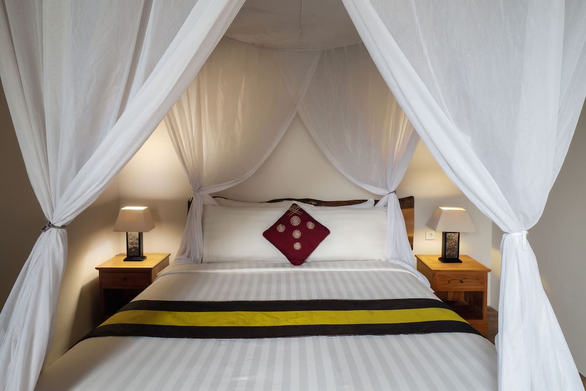 Una cama con colcha a rayas, un almohajarro rojo y un dosel. Hay dos mesitas de noche con lámparas.