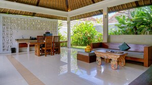 Una habitación amplia con techo alto, un área de comedor con muebles de madera, un sofá con cojines y un ventanal con vistas a zonas verdes.