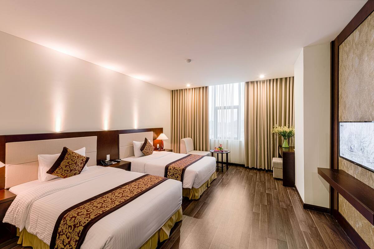 Ein Hotelzimmer mit zwei Betten, einem Schreibtisch, einem Stuhl, einem kleinen Tisch mit einer Vase und einem großen Fenster mit Vorhängen.