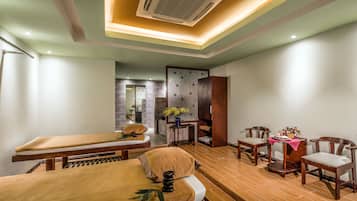 A spa room with two massage tables, wooden chairs, a small table with flowers, and a decorative wall.