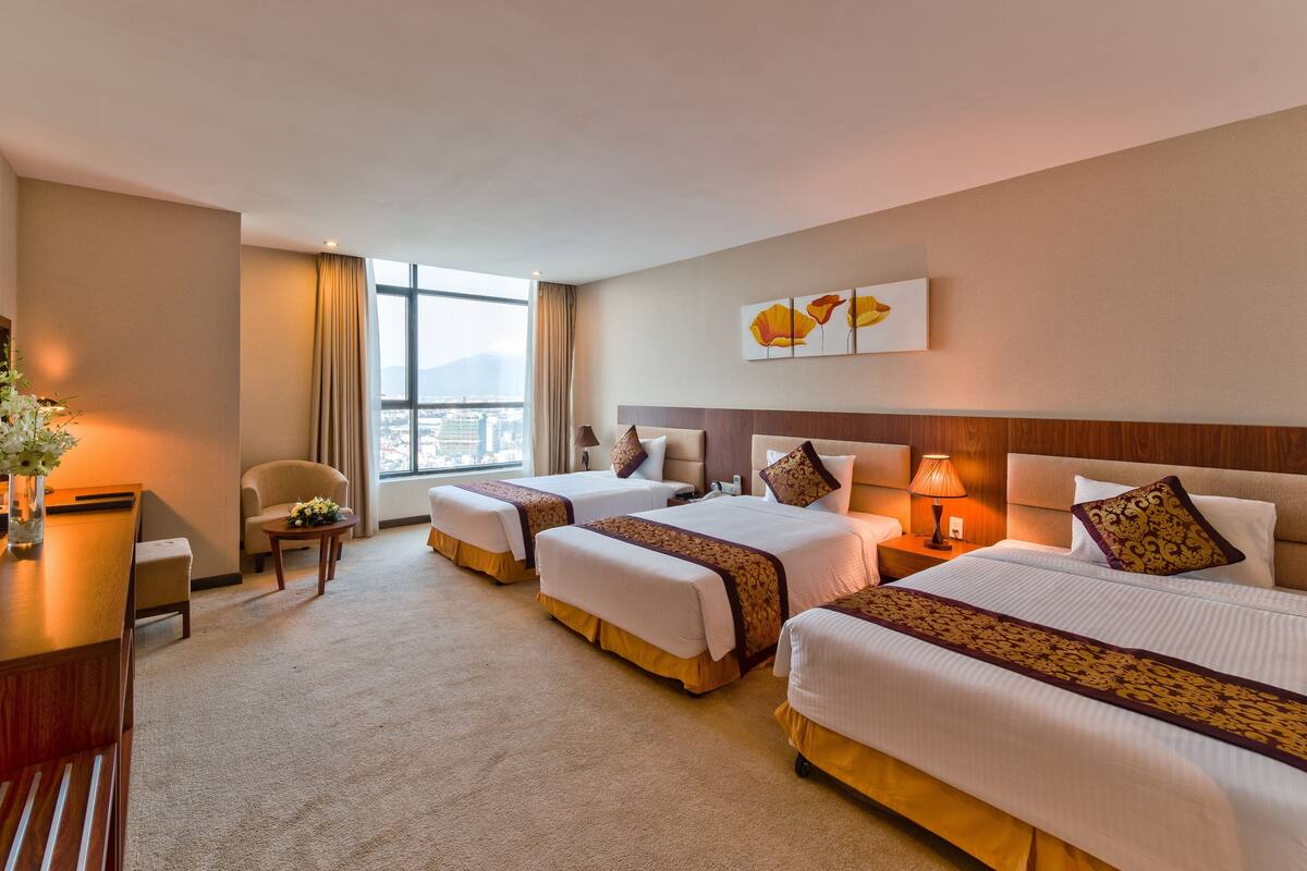 Ein Hotelzimmer mit drei Betten, einem Schreibtisch, einem Stuhl und einem großen Fenster mit Blick auf die Stadt.