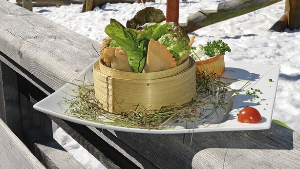 A wooden table with a basket of food, a plate with a tomato, and a mountain view in the background. - Venet Gipfelhütte (Zams)