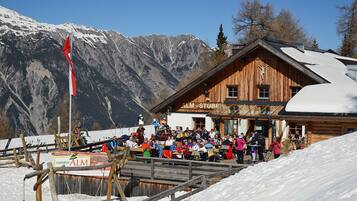 Eine Holzhütte in den Bergen, mit einem Schild mit der Aufschrift 'ALM-STUBE' und einer Gruppe von Personen, die draußen zusammengekommen sind.