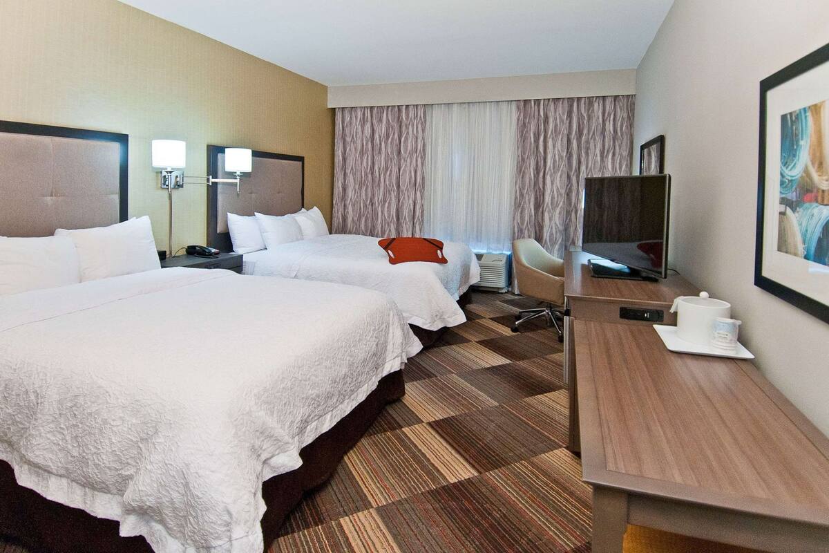 A hotel room with two beds, a desk, a chair, a TV, and a large window with curtains.
