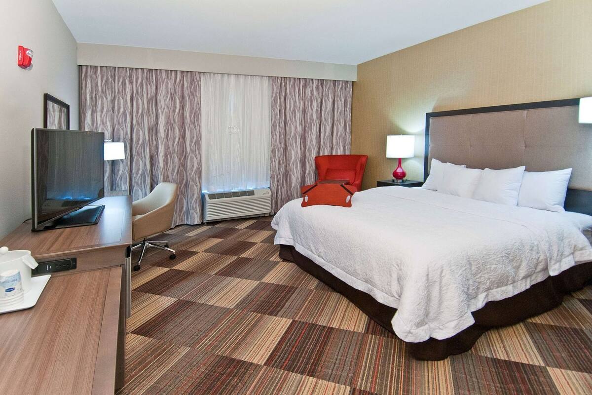 A hotel room with a bed, a desk with a laptop, a chair, a TV, and an air conditioning unit.