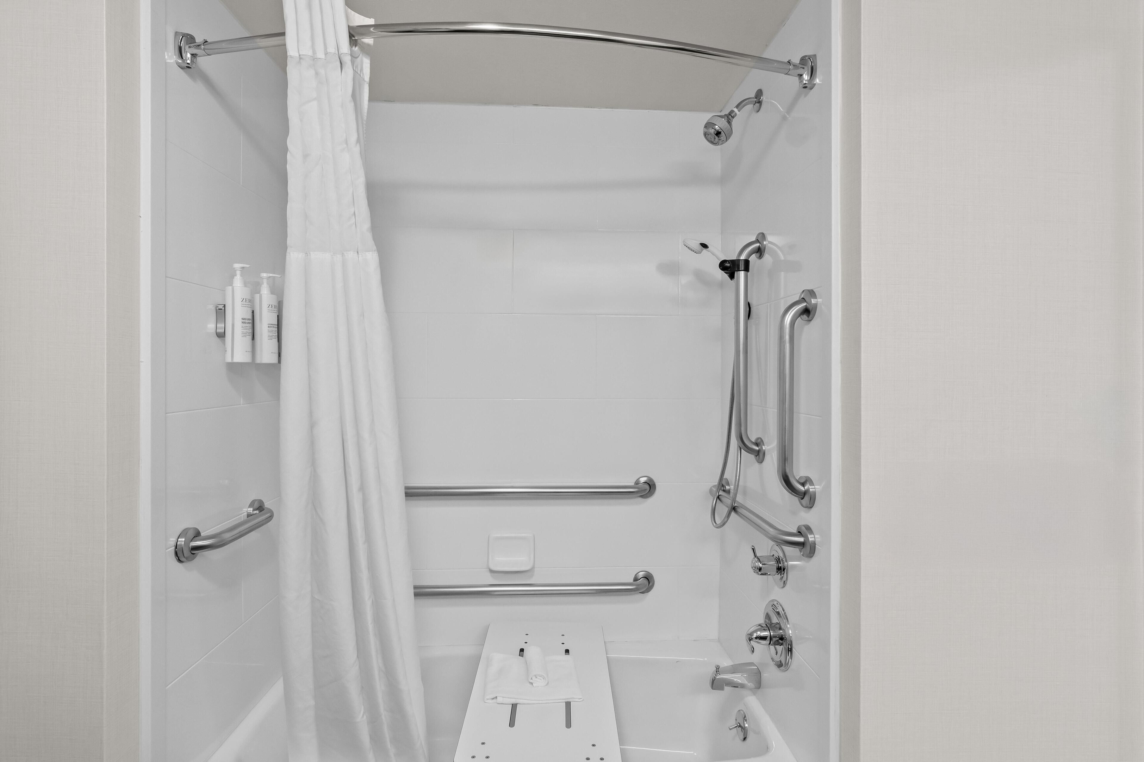 A bathroom with a shower, grab bars, a towel rack, and a soap dispenser.