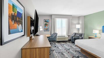 A hotel room with a bed, desk, TV, chairs, air conditioning unit, and artwork on the walls.