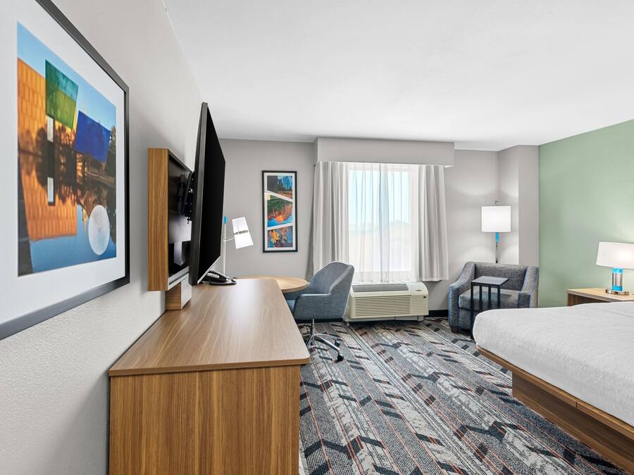 A hotel room with a bed, desk, TV, chairs, air conditioning unit, and artwork on the walls.