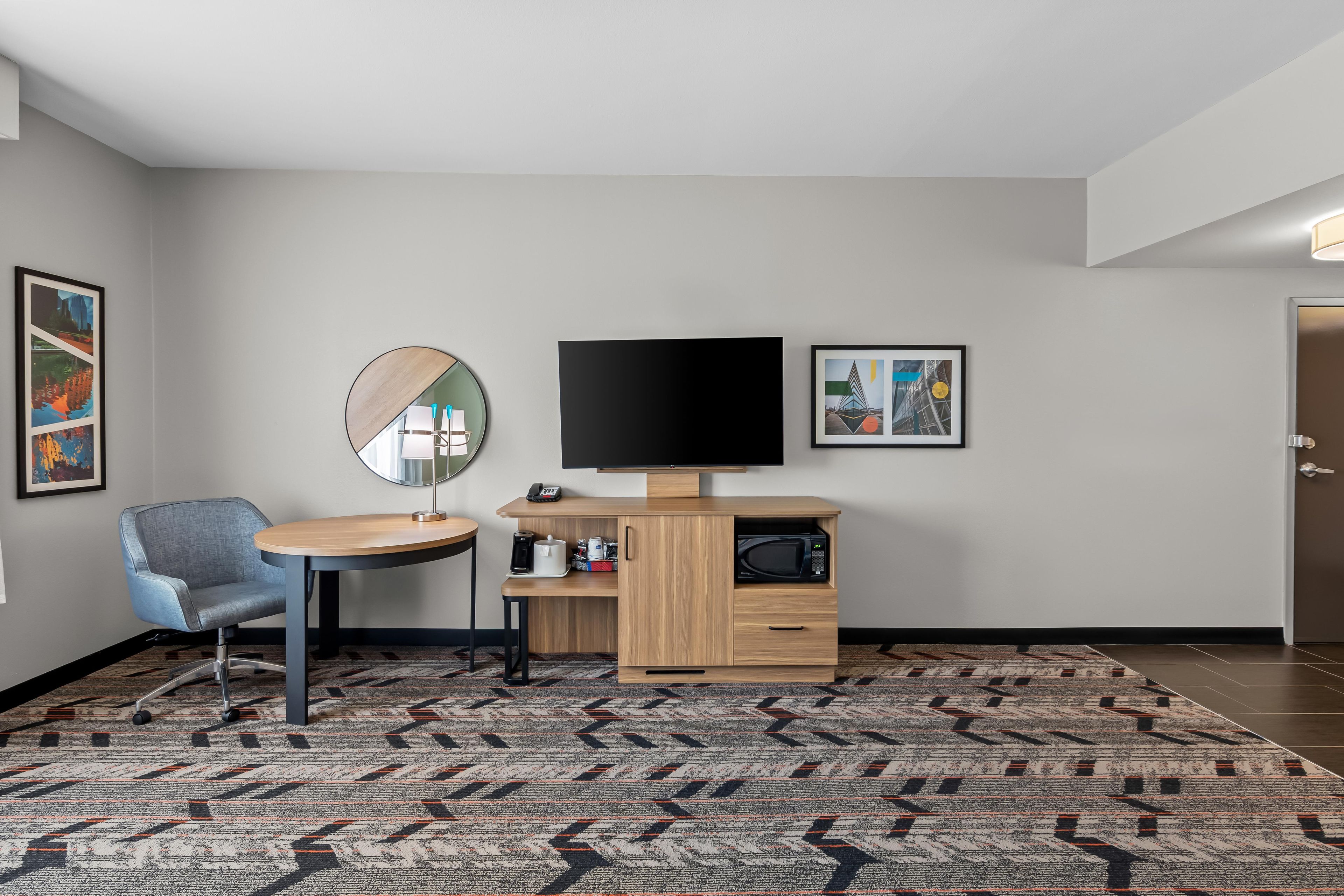 Habitación de hotel con una mesa redonda, una silla azul, un televisor de pantalla plana, un microondas, una cafetera y una alfombra estampada.