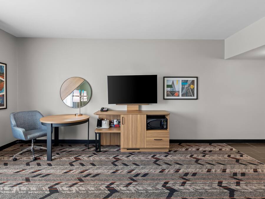 Habitación de hotel con una mesa redonda, una silla azul, un televisor de pantalla plana, un microondas, una cafetera y una alfombra estampada.