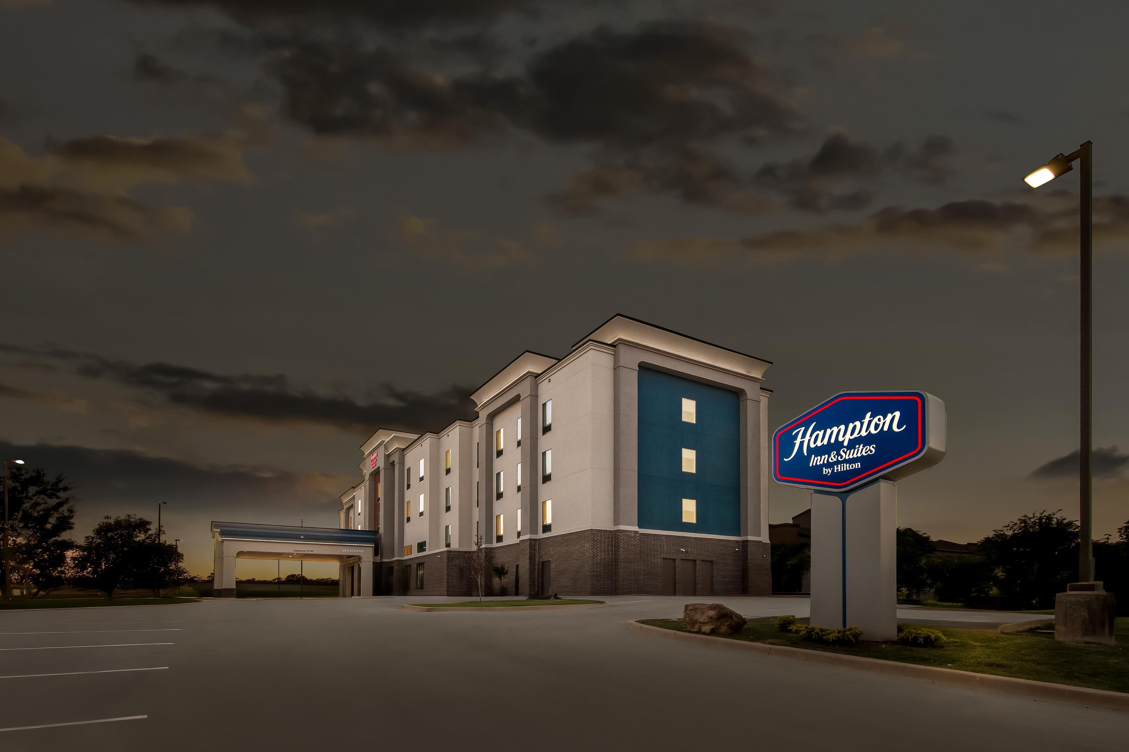 Edifici d'un Hampton Inn & Suites by Hilton, amb un gran cartell, un pàrquing i cel blau.