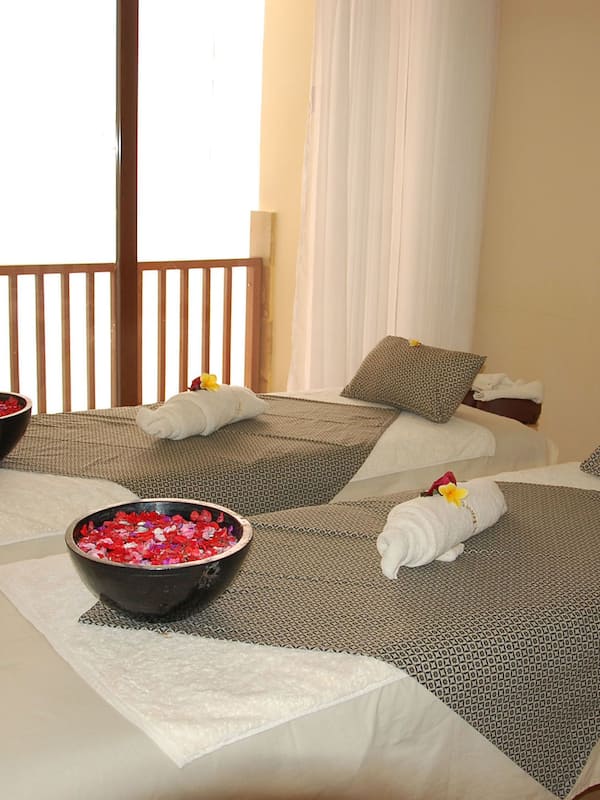Two plush beds, adorned with patterned bedspreads and pillows, invite relaxation. A serene ambiance is enhanced by a nearby bowl of rose petals and a beautiful painting on the wall.