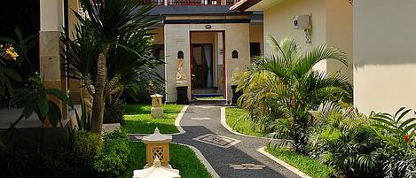 A courtyard with a pathway leading to a building entrance, surrounded by greenery and decorative elements.