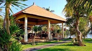 A covered outdoor area with tables and chairs, surrounded by palm trees and a view of the water. - Bali Dive Resort and Spa (Tulamben)