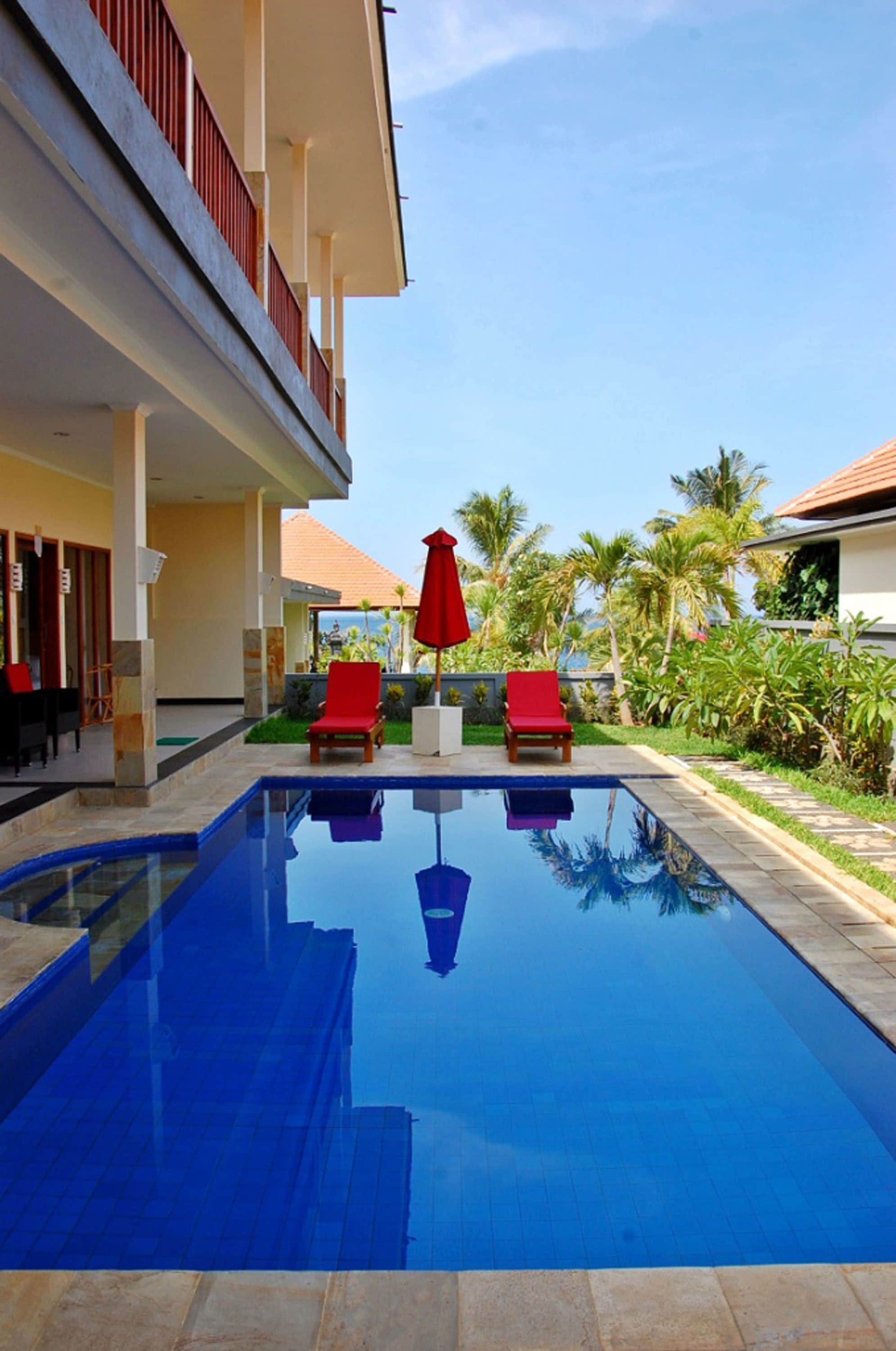 A swimming pool with red lounge chairs and umbrellas, surrounded by greenery and a building with balconies.