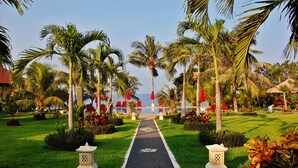 A pathway leading to a pool area surrounded by palm trees and red chairs, with a clear view of the sea in the background. - Bali Dive Resort and Spa (Tulamben)
