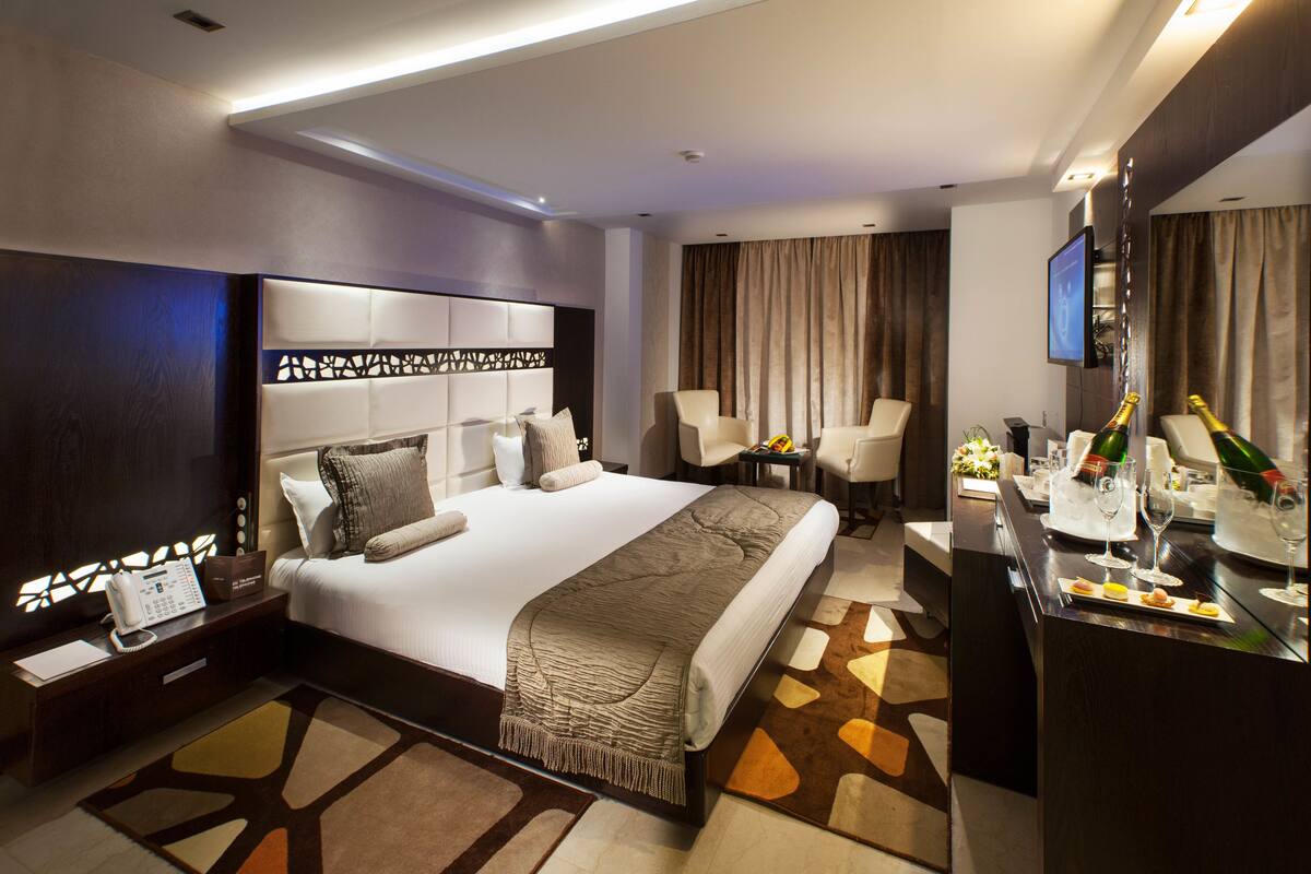A modern hotel room with a large bed, a desk with a computer, a TV, and a minibar.