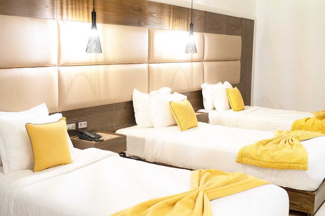 A hotel room with three beds, each with white bedding and yellow accents. There are bedside tables with lamps and a phone on one of them.