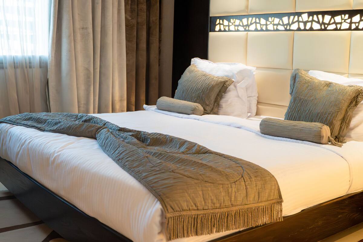 A neatly made bed with a brown throw blanket and pillows, a headboard with a decorative panel, and a window with curtains.