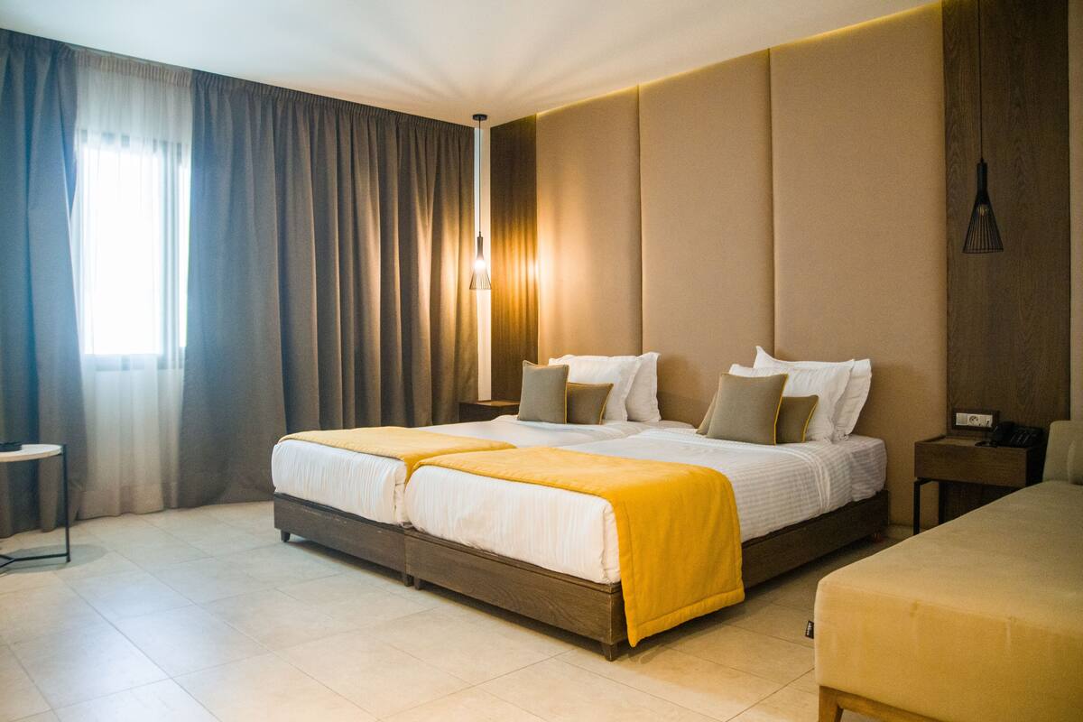 A hotel room with a double bed, a bedside table, a lamp, and a view of the outside through the window.