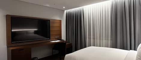 A hotel room with a bed, a desk with a television, and a large window with curtains.