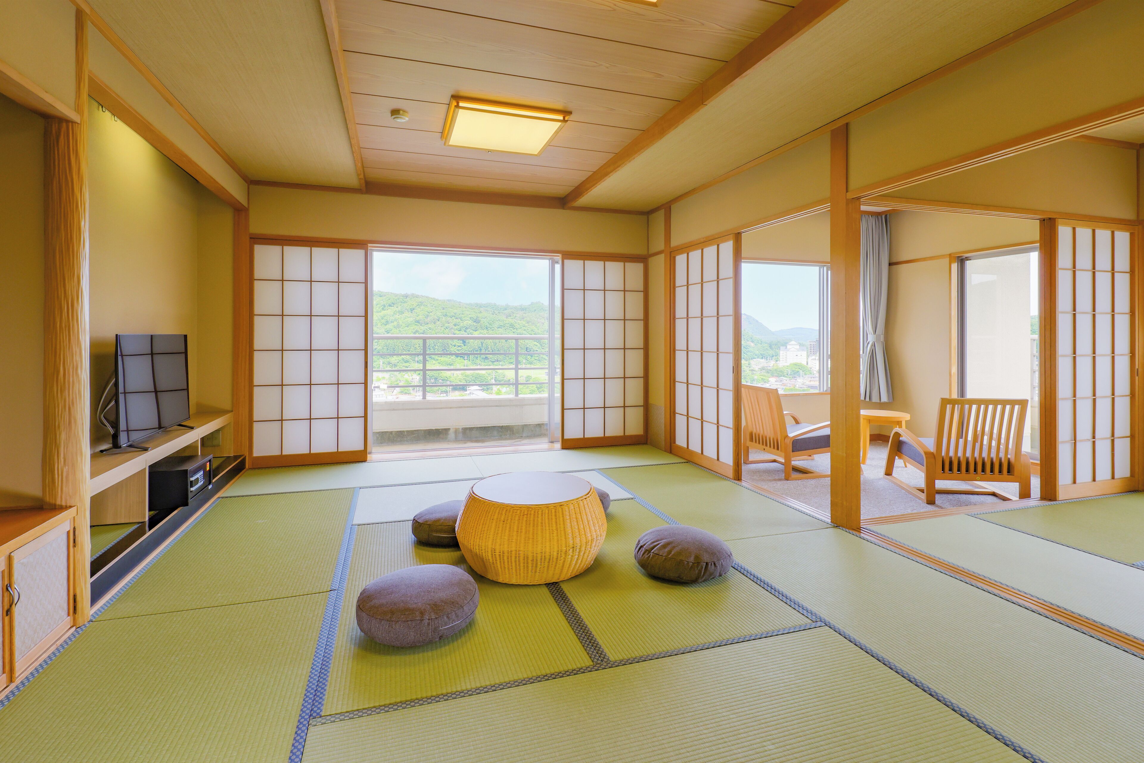 Una habitación japonesa tradicional con suelo de tatami, una mesa baja de tatami, un armario de madera bajo y una puerta corredera que da a un balcón con vistas a colinas verdes.