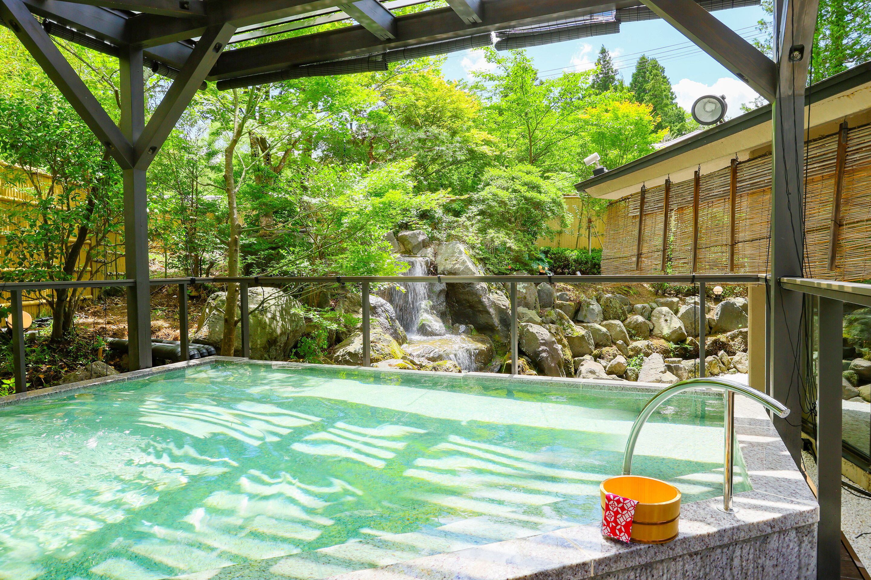 A hot tub with a waterfall backdrop, surrounded by greenery and a stone wall.
