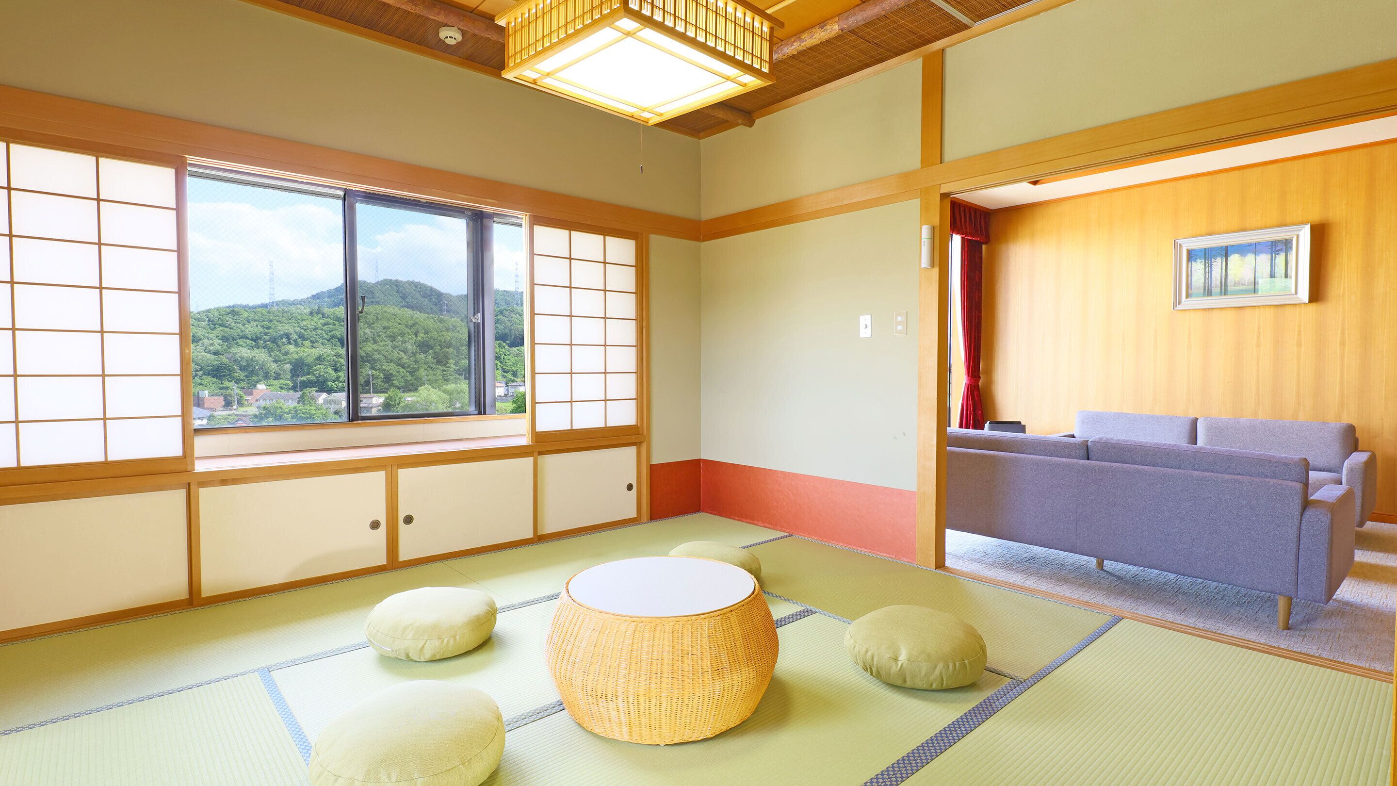A traditional Japanese room with tatami flooring, a tatami mat table, cushions, a sofa, and a view of a forested area through large windows.