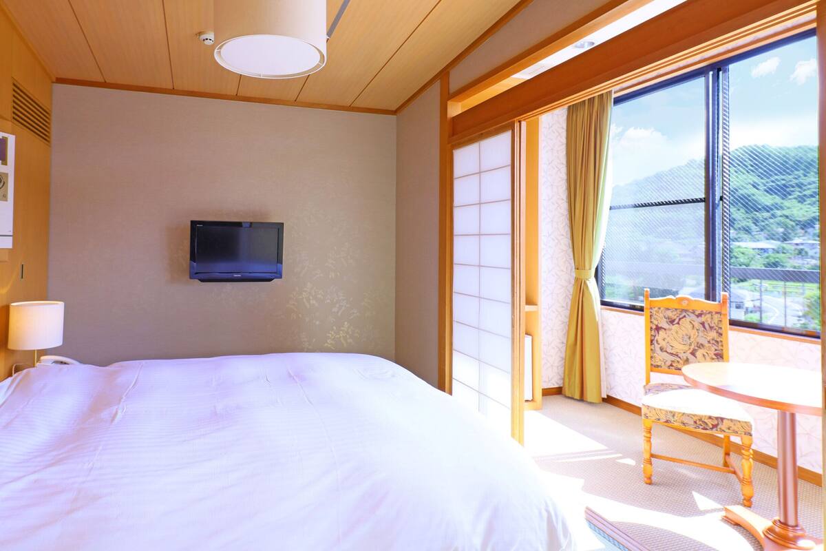 Une chambre d’hôtel avec un grand lit, un téléviseur fixé au mur, une chaise et une table près d’une grande fenêtre donnant sur un beau paysage, ainsi qu’un tableau d’affichage où sont accrochés divers objets.