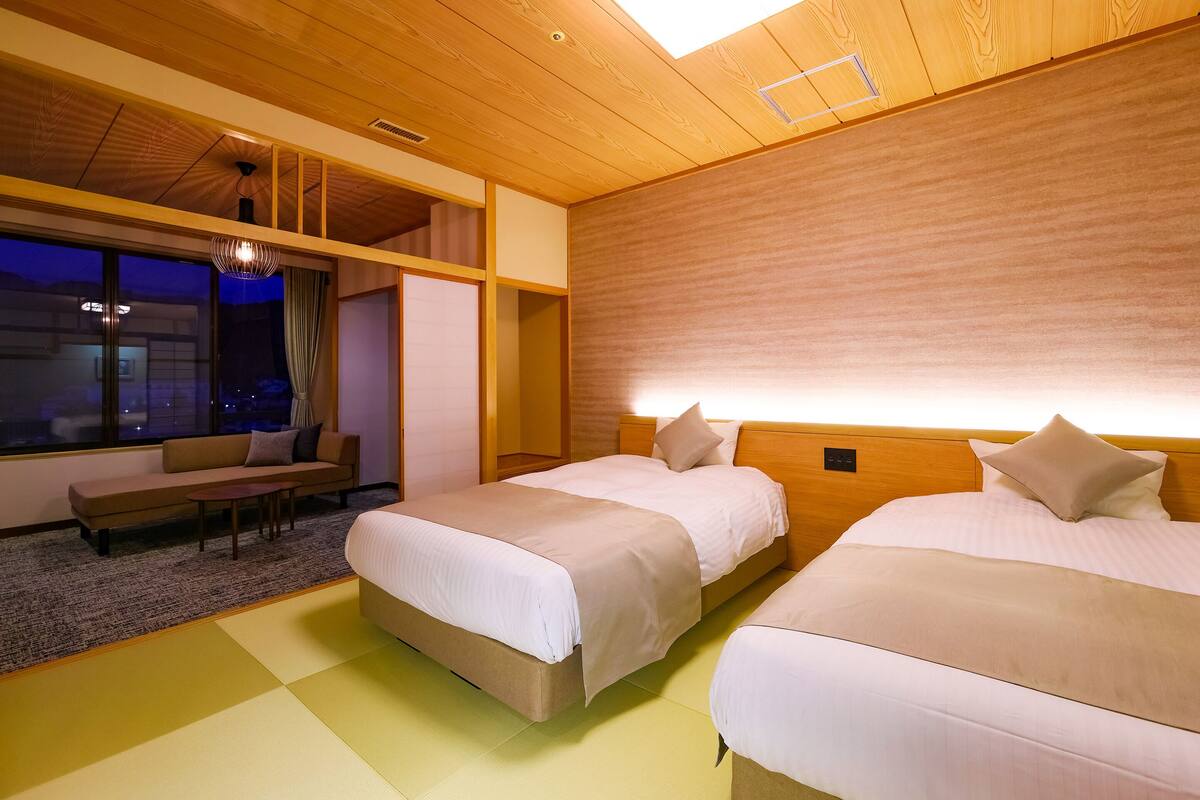 Une chambre d’hôtel avec deux lits, un canapé, une petite table et une grande fenêtre donnant sur la ville la nuit.