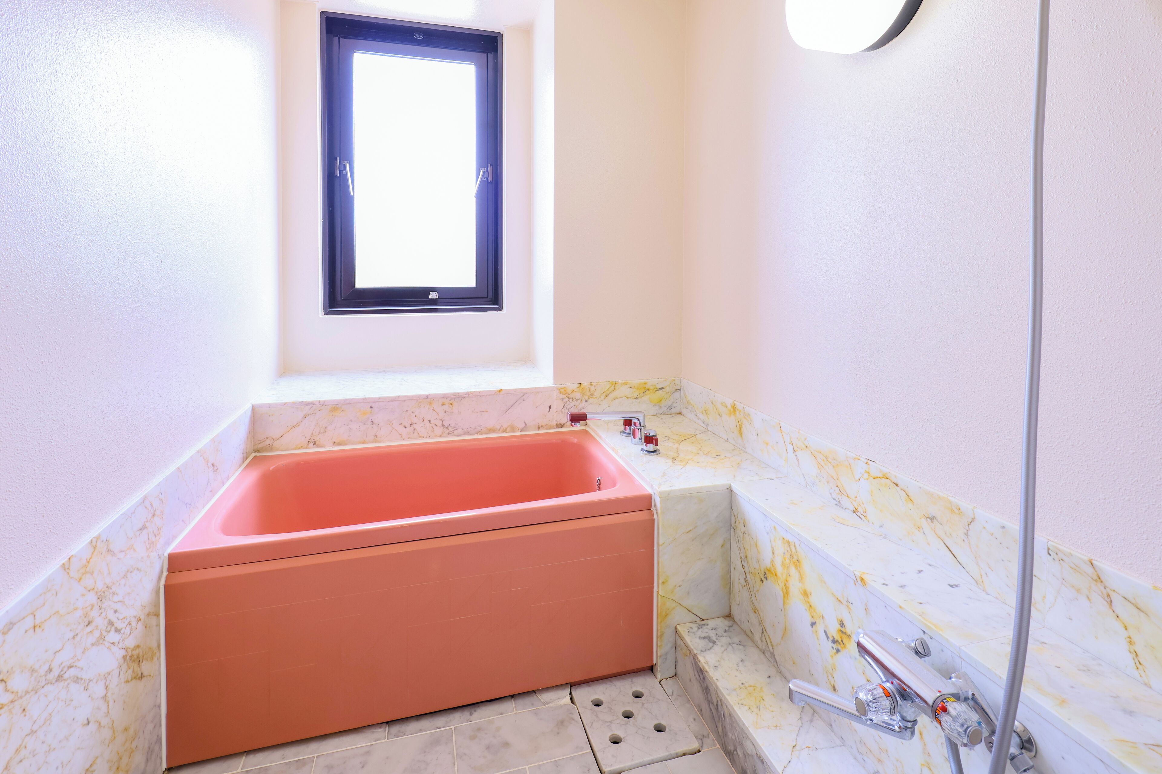 Een badkamer met een groot rechthoekig bad, een raam erboven en een wand betegeld met marmer.