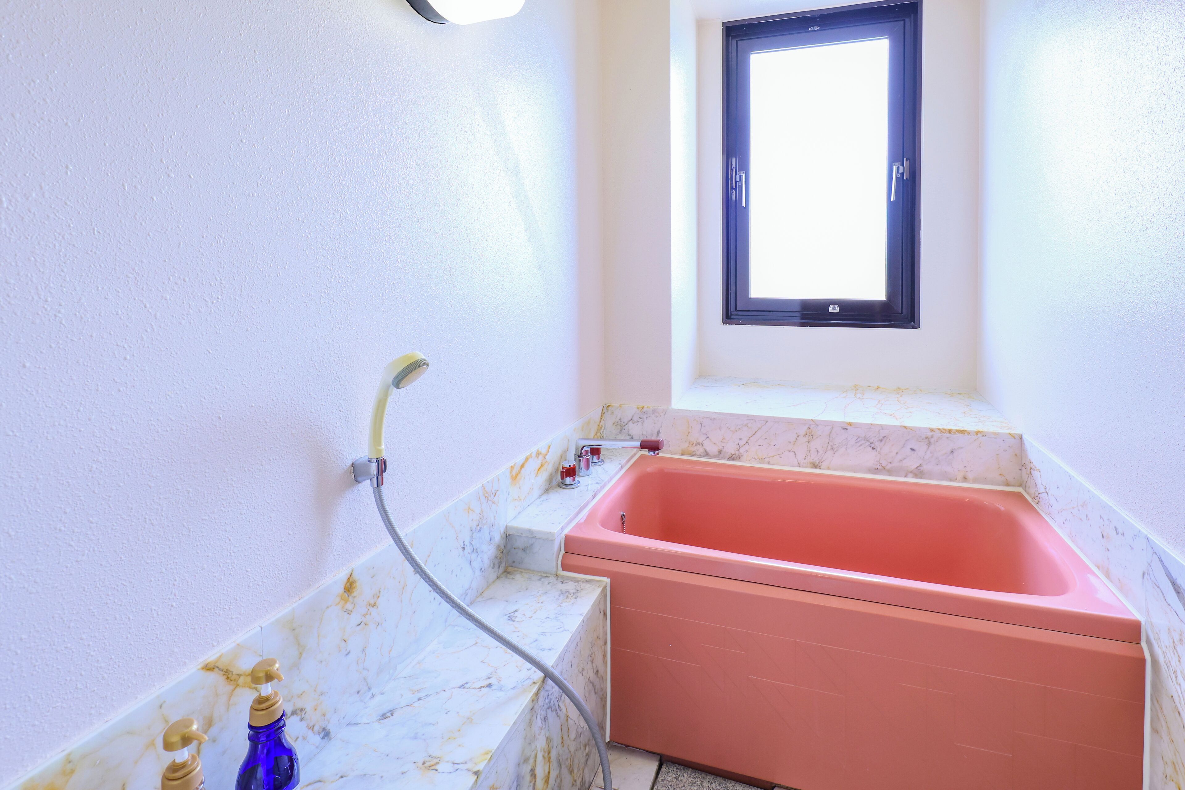 Un baño con una bañera rosa, una ventana y paredes de mármol.