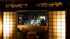 A traditional Japanese room with tatami flooring, a low wooden table, and two chairs. A large window offers a view of a cityscape at night. - Hotel Takasago (Kochi)