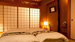 A traditional Japanese-style room with tatami flooring, a low wooden bed with white bedding, a shoji screen, and a bedside lamp with a vase of flowers. - Hotel Takasago (Kochi)