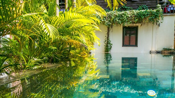 A clear pool with reflections of palm trees, a white building with a Coca-Cola sign, and a window with a red curtain. - River and Villa (Siem Reap)