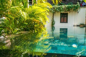 A clear pool with reflections of palm trees, a white building with a Coca-Cola sign, and a window with a red curtain. - River and Villa (Siem Reap)