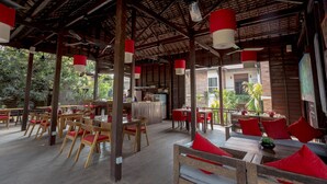An open-plan restaurant with wooden furniture and red cushions, featuring a high ceiling with exposed beams and hanging red and white pendant lights. - River and Villa (Siem Reap)