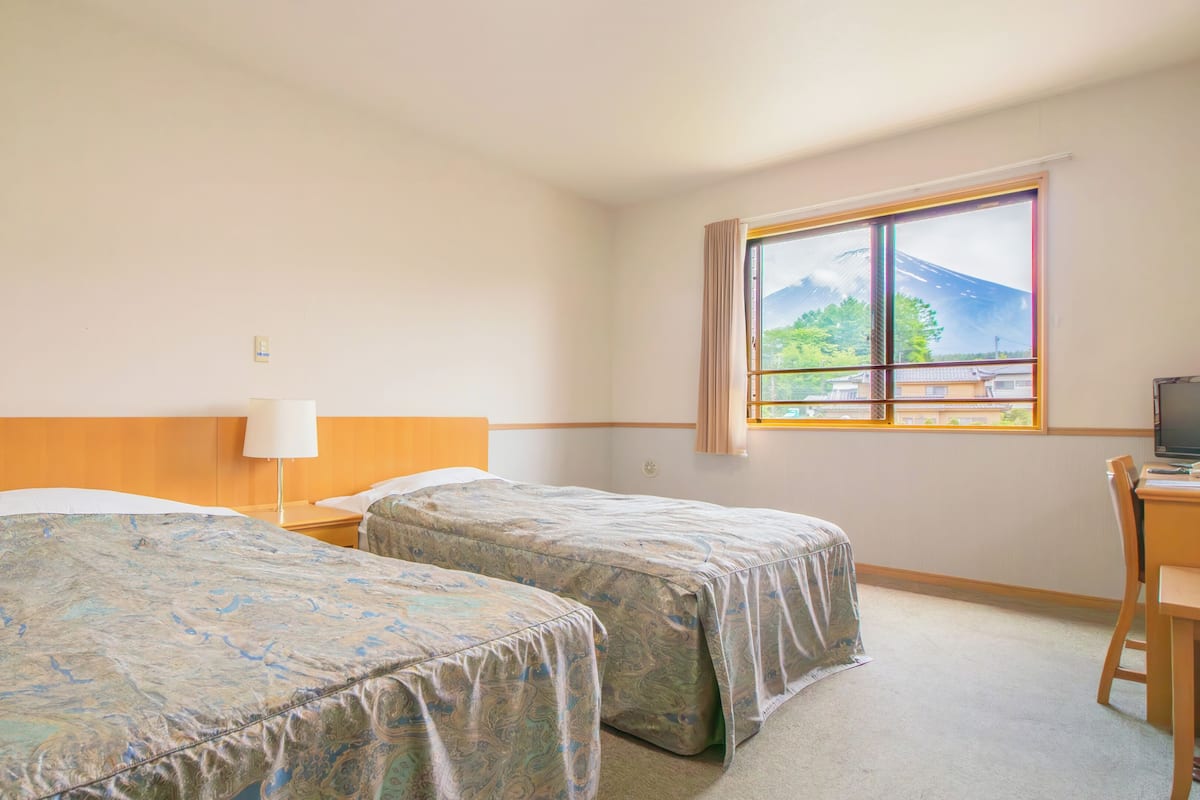 A hotel room with two beds, a desk, a chair, a TV, and a window with a view of a mountain and buildings.
