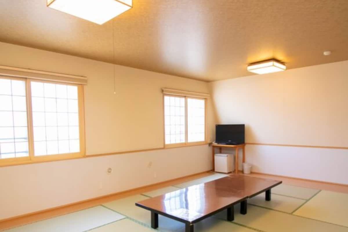 A room with tatami flooring, a low table, a television on a stand, and large windows.