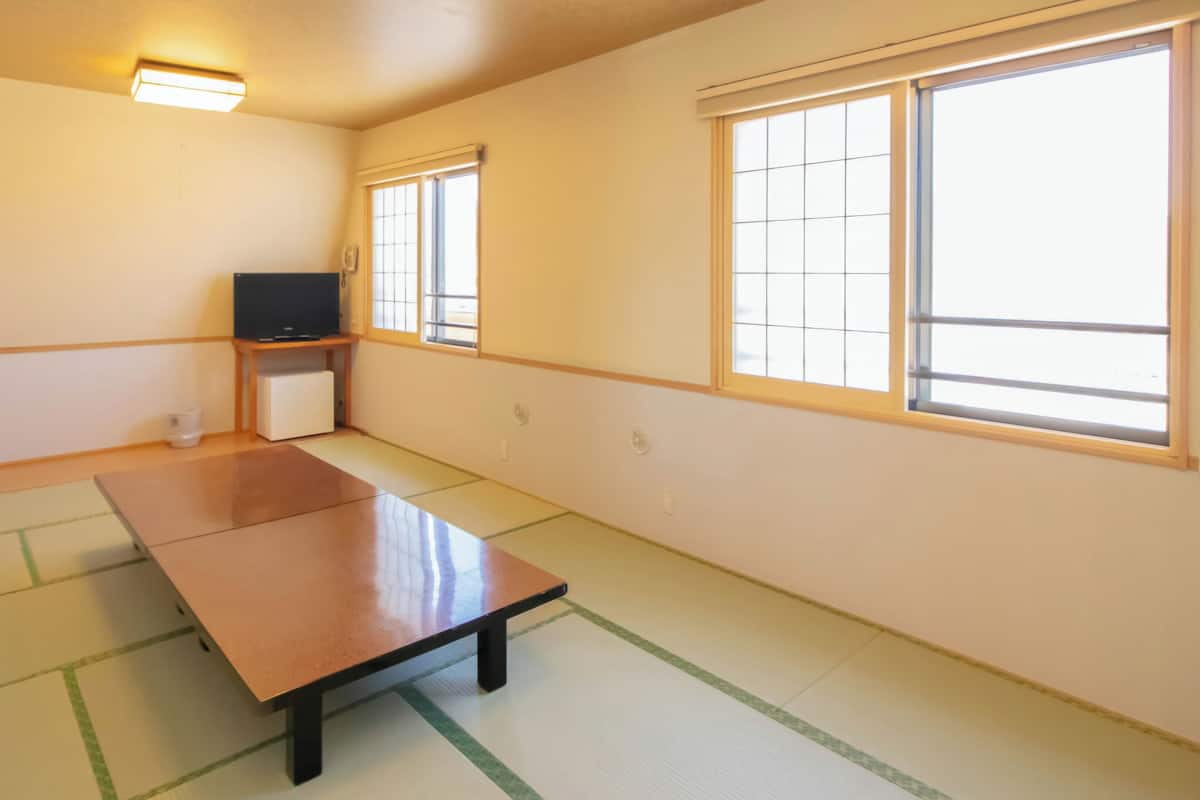 A room with tatami flooring, a low table, a television on a stand, and large windows.