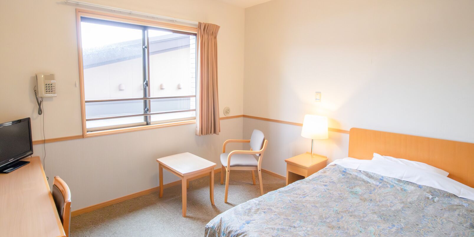 A hotel room with a bed, a desk, a chair, a TV, a window with curtains, and a lamp.